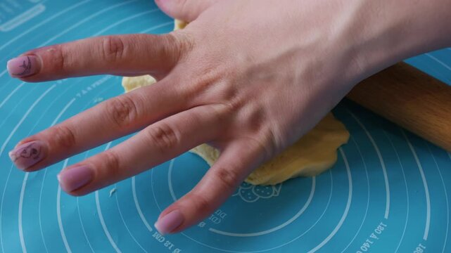 Hands use a wooden rolling pin to flatten fresh yeast dough on a blue silicone pastry mat with circular measurements. Concept of home baking, culinary skills, and pastry preparation.