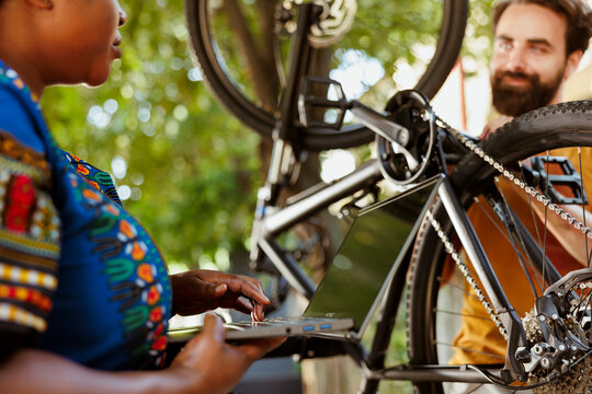 Closeup shot of black woman searching on minicomputer to help young man fix damaged bike. Sporty boyfriend and girlfriend servicing bicycle chain while using laptop for annual summer outdoor cycling.