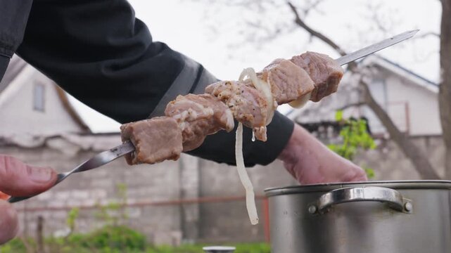 Male hands with vitiligo string pieces of raw marinated meat and onions onto a metal skewer. The person prepares food for an outdoor barbecue meal next to a metal pot.