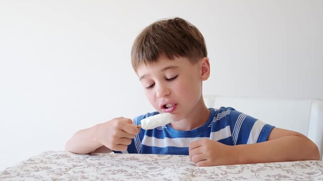 Handsome boy eating popsicle on a stick. He bites off the ice cream with pleasure. Close-up. Child eat in the kitchen or at the dining room table. Tasty dessert. Slow motion. High quality 4k footage.