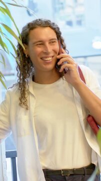 Vertical video: Answering phone call, adult in white tee fixing red strap bag at glass door, plant