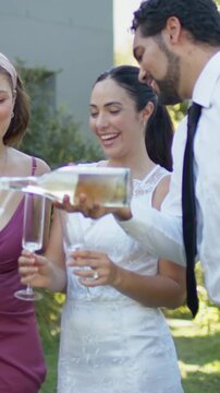 Vertical video: Tilting groom pouring bottle over flutes in garden, bride in white lace celebrating