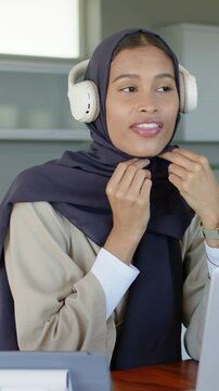 Vertical video: Woman hearing laptop ping adjusting hijab, donning white headset speaking for call