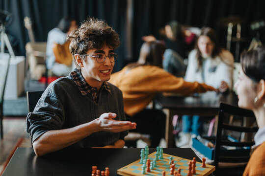 A young male student gestures while talking to a fellow student over a chessboard. Other students socialize and play games in the background of a casual study or club space.
