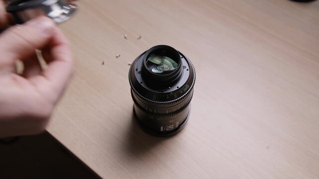 Hands carefully placing and fitting a PL bayonet mount onto a cinema lens. Final step of an EF to PL mount conversion. Precise lens modification process shown on a wooden desk.