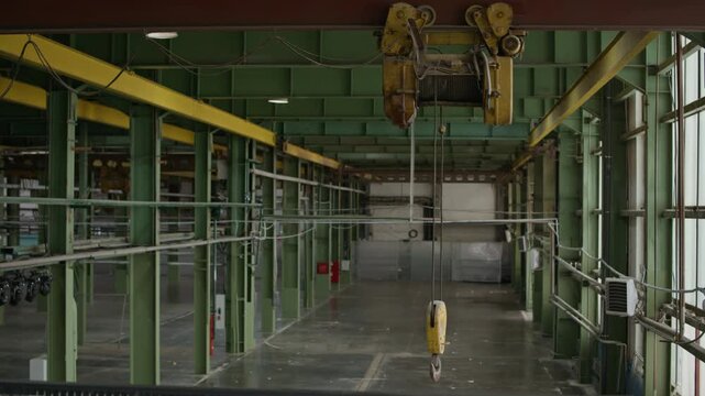 Wide establishing shot of spacious empty industrial warehouse featuring green steel columns, overhead crane hoist system and large open floor area