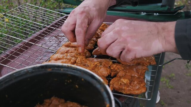 A man with vitiligo skin patches carefully arranges marinated chicken pieces on a metal grill grate. The meat is coated in a vibrant orange marinade for an outdoor barbecue.