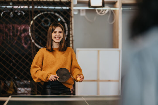 A smiling young woman plays table tennis holding a paddle and ball. She enjoys a casual indoor game while a friend watches nearby.