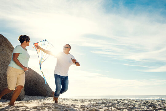 Mature, couple and flying with kite on beach for summer holiday, playful bonding or vacation. Man, woman or playing with wind or blue sky on ocean coast for weekend trip or freedom on mockup space