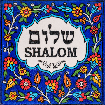 Colorful Armenian tile that says Shalom in English and Hebrew text on white with a flowered blue background.
