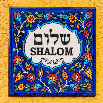 Colorful Armenian tile that says Shalom in English and Hebrew text on white with a flowered blue background against a yellow wall.
