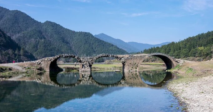 Close-up time-lapse of the ancient three-arch stone bridge with blue sky, clear water, and mirror-like reflections in spring, Wuyuan county, Jiangxi province, China.