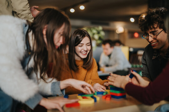 A group of students and friends play with colorful wooden blocks and smile together. The casual indoor scene shows young adults enjoying a cooperative tabletop game and social time.