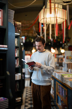 Relaxed thoughtful man visiting popular book fair in local readers market and choosing books to purchase. Pondering male bookworm holding book and reading content of novel in trendy bookstore.