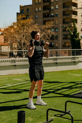 Fototapeta premium Fit young man lifting dumbbells overhead during strength workout in outdoor gym. Fitness training, power and endurance concept.