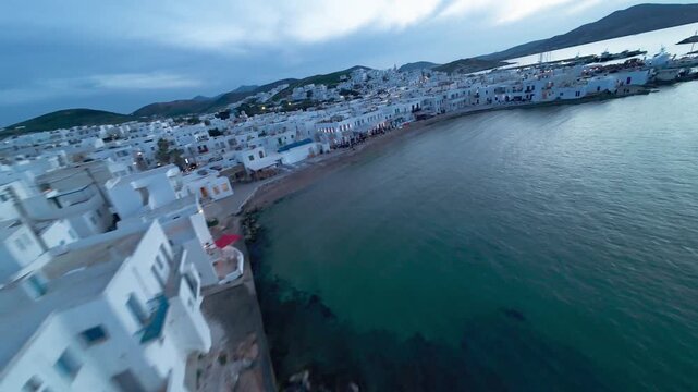 Cinematic FPV drone flight over Naousa village on Paros island during sunset, featuring warm evening light, coastal harbor and Easter celebrations with festive atmosphere