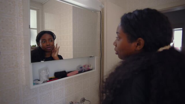 African American woman applying skincare while looking in bathroom mirror during morning routine creating calm personal care moment in home interior