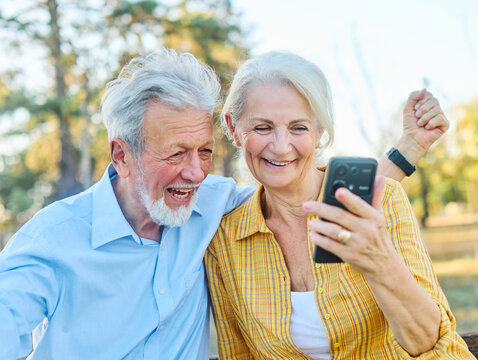 Happy active senior couple taking selfie sitting ob a bench in park outdoors. Love togetherness and acive seniors vitality concept