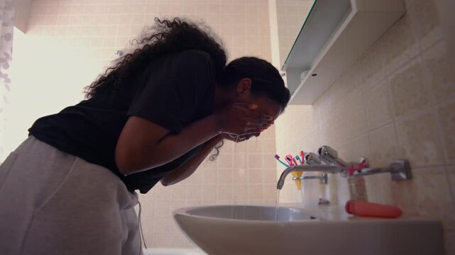 African American woman washing face over sink with running water during daily hygiene routine creating refreshing and focused personal care moment at home