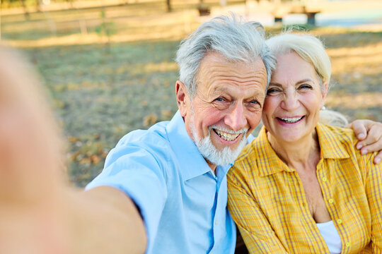 Happy active senior couple taking selfie sitting ob a bench in park outdoors. Love togetherness and acive seniors vitality concept