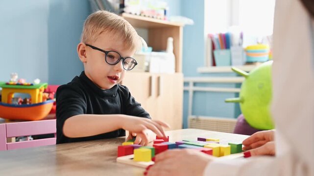 Young boy wearing glasses plays with colorful building blocks on a wooden table, while a teacher's hand assists him in a bright classroom environment