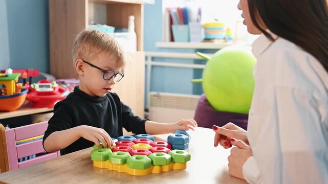 Young boy with glasses plays with colorful blocks while seated at a table with a teacher in a vibrant classroom filled with toys and learning resources