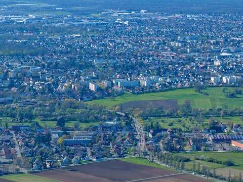 Aerial view of sprawling cityscape meeting verdant fields under a soft, diffused light, casting long shadows across the landscape, Sausheim, Grand Est, France.