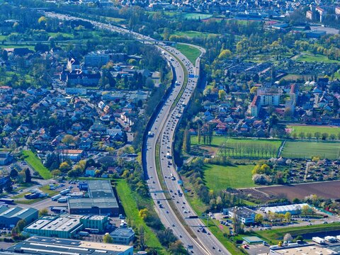 Aerial view of a serpentine highway cutting through urban sprawl, bordered by vibrant green fields and dense trees, Sausheim, Grand Est, France.