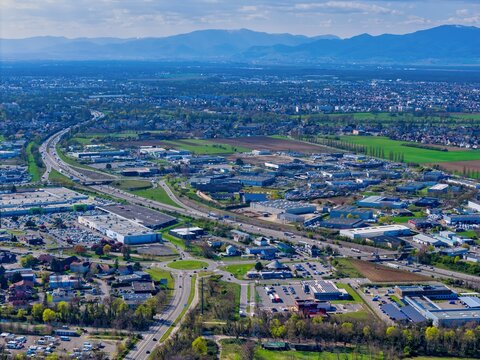Aerial view of sprawling urban landscape, highways cutting through the city, mountains standing tall in the distance, a symphony of progress and nature, Sausheim, Grand Est, France.
