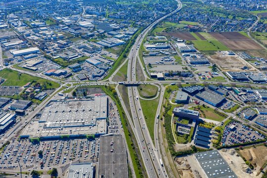 Aerial view of a highway cutting through a city, surrounded by industrial buildings and green fields, creating a stark contrast between urban development and nature, Sausheim, Grand Est, France.