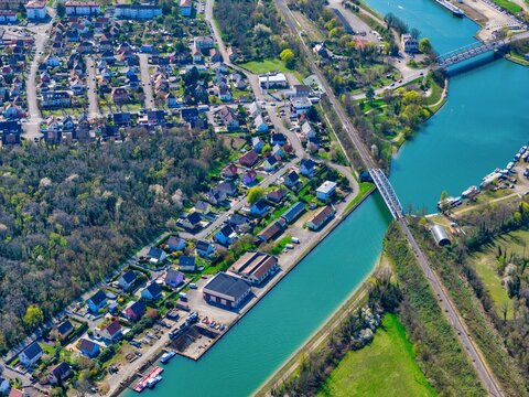 Aerial view of the tranquil canal reflecting the sky's soft hues, contrasting with the terracotta roofs and the verdant trees near the houses, Sausheim, Grand Est, France.