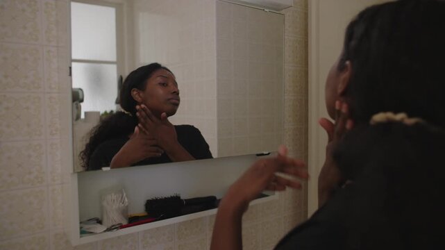 African American woman spreading moisturizing cream across face while looking at her reflection in bathroom mirror creating calm and attentive skincare moment in home interior
