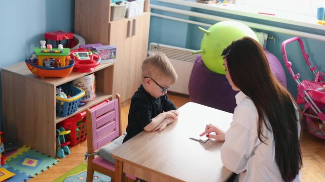Young boy wearing glasses participates in educational activity with teacher, colorful learning cards spread on the table in a bright classroom environment