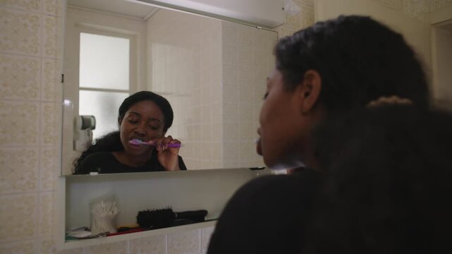 African American woman brushing teeth in front of mirror with focused expression during daily hygiene routine creating steady and quiet personal care moment at home