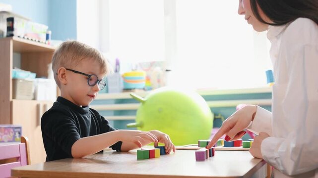 Young boy wearing glasses plays with colorful building blocks on a wooden table, while a teacher's hand assists him in a bright classroom environment