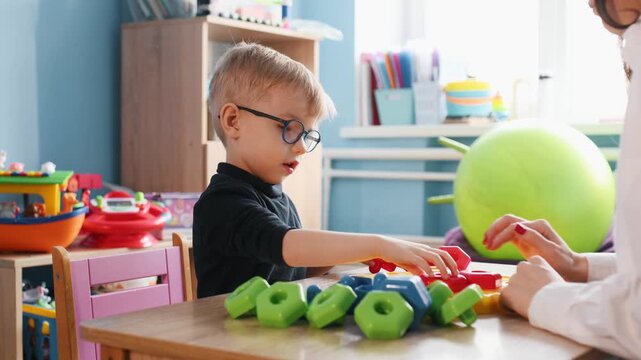 Young boy wearing glasses plays with colorful building blocks on a wooden table, while a teacher's hand assists him in a bright classroom environment