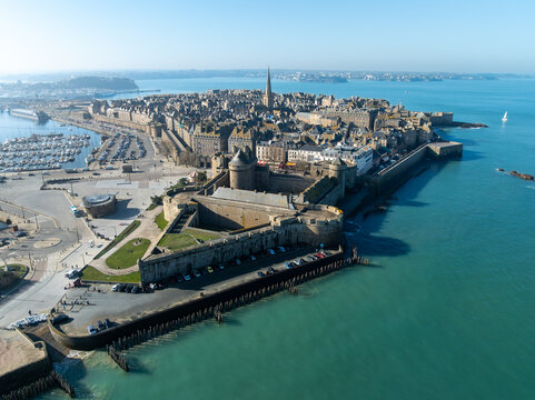 Aerial view of the ancient walled city, its ramparts kissed by the turquoise sea, with the spire of Saint-Malo Cathedral piercing the clear blue sky, Saint-Malo, Brittany, France.