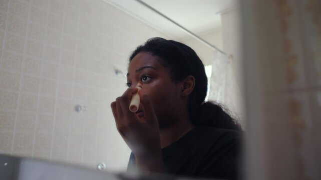 African American woman blending makeup on cheek with brush then tapping product with finger while looking at her reflection in bathroom mirror creating detailed beauty routine moment at home