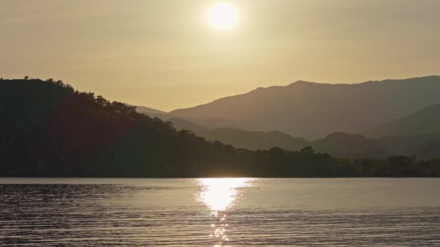 Scenic sunset over a tranquil Phaselis Bay with golden sun reflection on calm water and silhouetted hills in the background - Aerial Parallax Shot