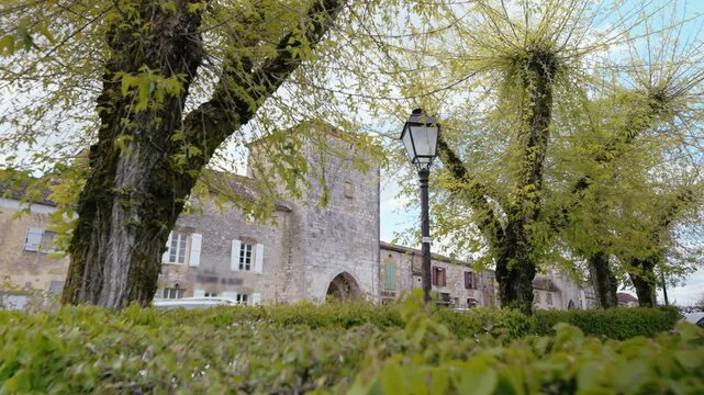 Medieval stone gateway and traditional houses in Monpazier, Dordogne, framed by green trees in a public square. Slow motion tracking shot.