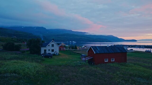 Kokelv, Hammerfest, Finnmark, Norway - A Peaceful Village Sits by the Sea, With Houses and a Red Barn Beneath Soft Pink Clouds and Blue Mountains - Aerial Drone Shot