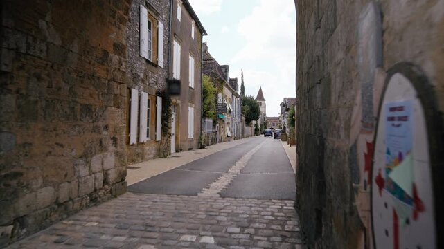 Historic stone houses line a narrow paved street leading to a distant church in Monpazier, Dordogne. Slow motion traveling shot at low height.