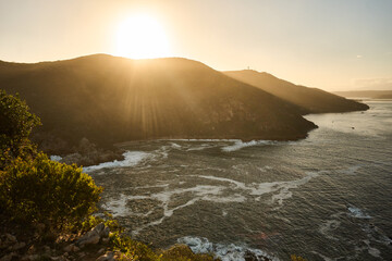 Obraz premium Sunlit Headland Overlooking Turbulent Ocean Waves, Bright Backlight And Crashing Surf, Rugged Hills