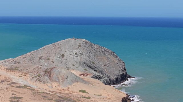 Drone footage capturing the vibrant blue sea and rugged landscape surrounding Pilon de Azucar, Cabo de la Vela, Colombia, during a clear, sunny day, showcasing nature's beauty.
