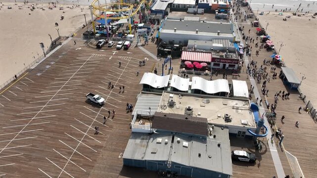 Aerial view of Santa Monica Pier in Santa Monica, California, facing Pacific Coast Highway with tourists walking along the pier and riding the Ferris wheel and roller coaster on a cloudy day.