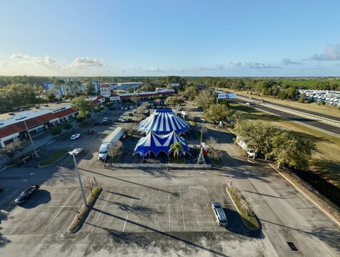 Aerial view of a vibrant blue and white circus tent standing proudly amidst a sprawling parking lot, framed by lush greenery and distant buildings, St. Augustine, Florida, United States.