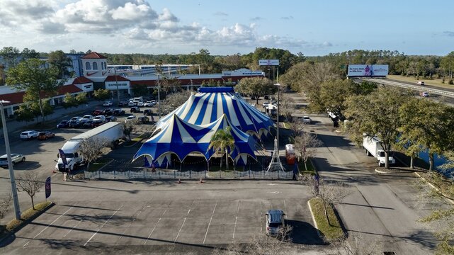 Aerial view of the grand blue and white striped circus tent standing proudly amidst a sea of parked cars, framed by the distant shops and highway, St. Augustine, Florida, United States.