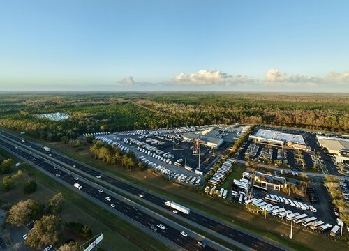 Aerial view of an RV dealership lot with rows of white RVs contrasting against the surrounding green forest and a multi-lane highway, St. Augustine, Florida, United States.