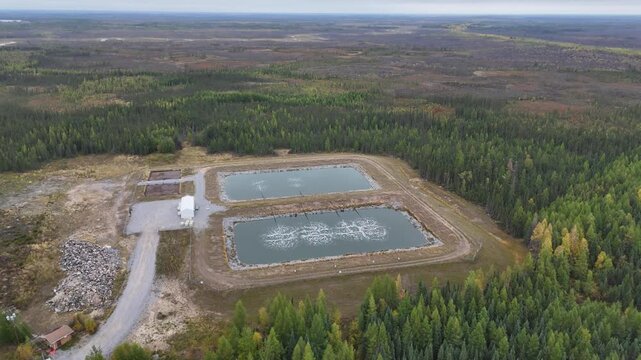 Aerial drone orbits left in a wide shot over wastewater lagoons near Ilford War Lake, Northern Manitoba, Canada. Aeration bubbles, berms, gravel road, and boreal forest visible from distance.