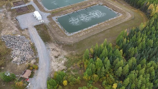 Aerial drone flies forward top-down over boreal forest, then pans up to reveal wastewater lagoons near Ilford War Lake, northern Manitoba, Canada. Aeration bubbles, berms, and gravel road visible.